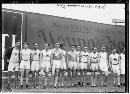 Lineup of young male athletes in sleeveless shirts and shorts posing outdoors in front of a wall with banner advertising behind them, early 20th-century photo