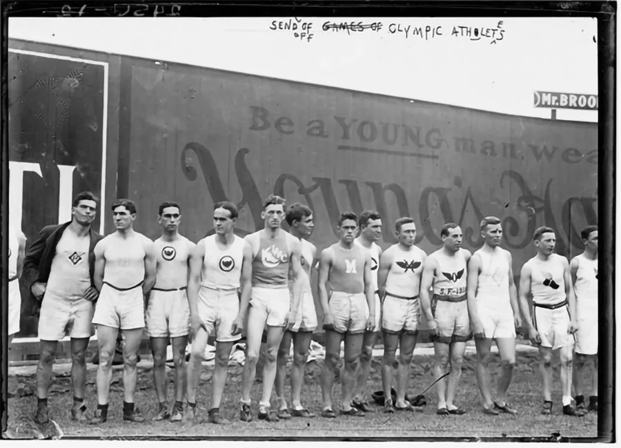 Lineup of young male athletes in sleeveless shirts and shorts posing outdoors in front of a wall with banner advertising behind them, early 20th-century photo