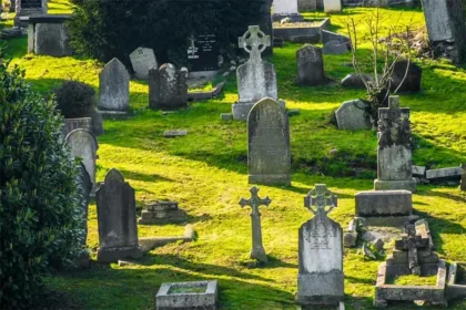 Old cemetery with weathered gravestones and a central Celtic cross, sunlit on green grass.