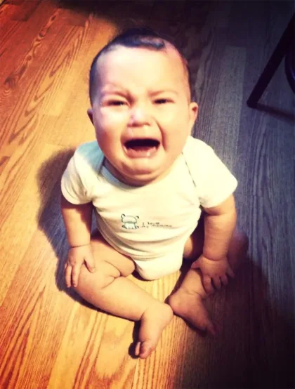 image_desktop 7 | Axxcess Platform Baby sitting on a wooden floor crying, wearing a white onesie with a small bear logo near the chest.