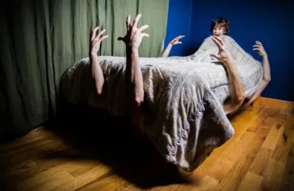 A staged horror scene: a person lies under a bed quilt, with several arms reaching up from beneath the covers in a dimly lit room.