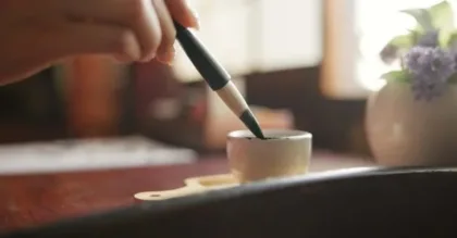 Person using a brush to glaze a small ceramic cup on a wooden table with a blurred background.