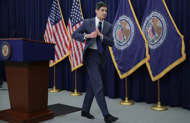 image_desktop 17 | Axxcess Platform Man in a dark suit walking past a blue podium, with American flags and government seals in the background at a press briefing setting.