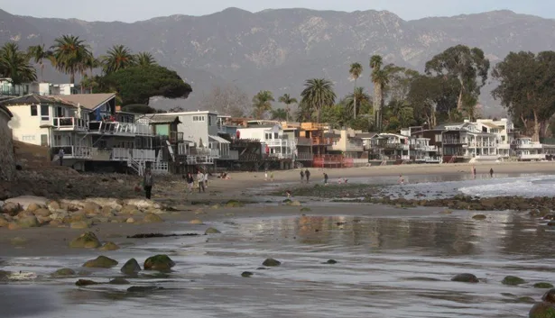 image_desktop 2 | Axxcess Platform Coastal village with beachfront houses, palm trees, and distant mountains along a reflective shoreline with people walking nearby.