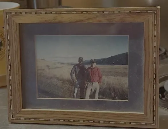 Framed photo of two men standing together in a grassy field with hills in the background.