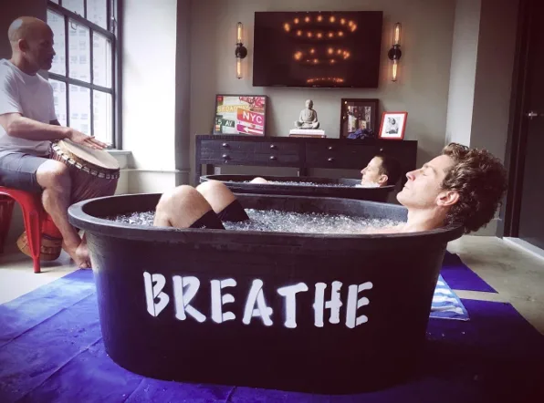 image_desktop 3 | Axxcess Platform Man reclining in a large black tub labeled 'BREATHE' while another person plays a tambourine nearby, in a modern wellness room with a Buddha statue and framed photos.