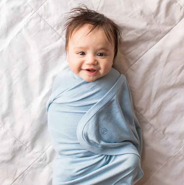 image_desktop 30 | Axxcess Platform Smiling baby wrapped in a light blue swaddle on a white quilted bed.