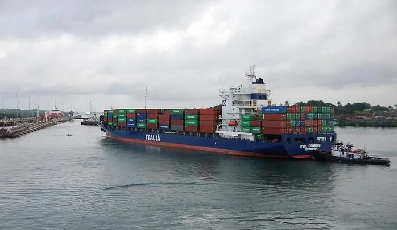 Large blue container ship Italia in a harbor, stacked with colorful shipping containers, aided by a tugboat at its bow.