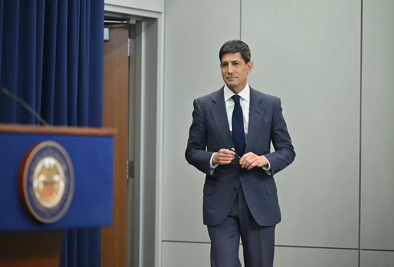 A man in a navy suit and tie stands beside a blue podium in a conference room, adjusting his glasses as he prepares to speak.