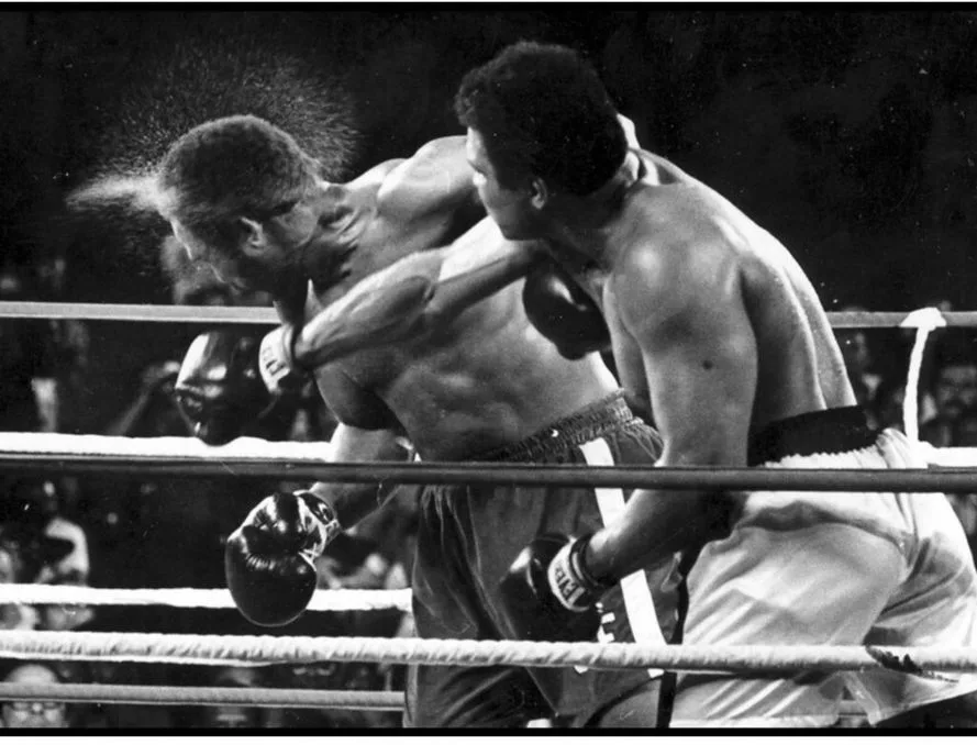 Two male boxers in a ring clash mid-fight as one lands a punch on the other.