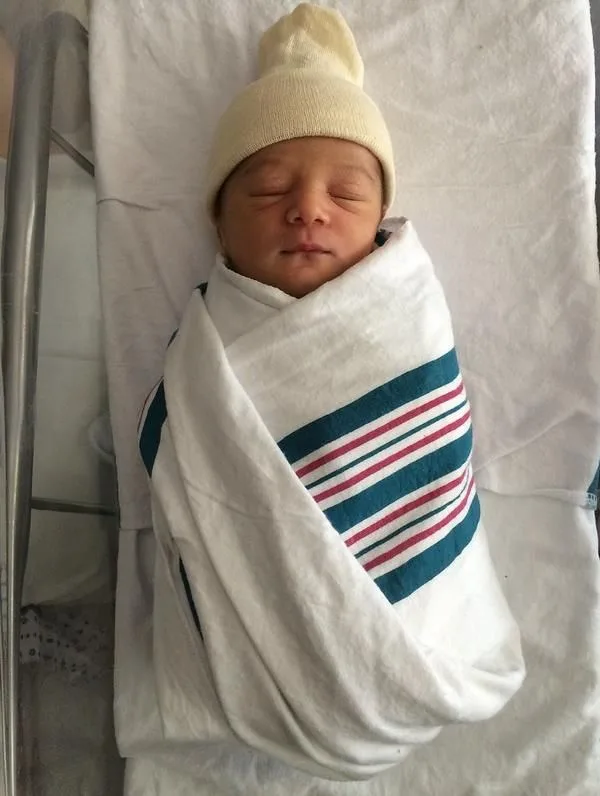 Newborn baby swaddled in a white blanket with teal and pink stripes, wearing a cream knit cap, lying in a hospital crib.